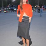 Victoria Hamuche wears a Chanel orange ribbed knit cardigan, a black midi dress with a white waist and train, black and white toe pumps, and red flower earrings.
