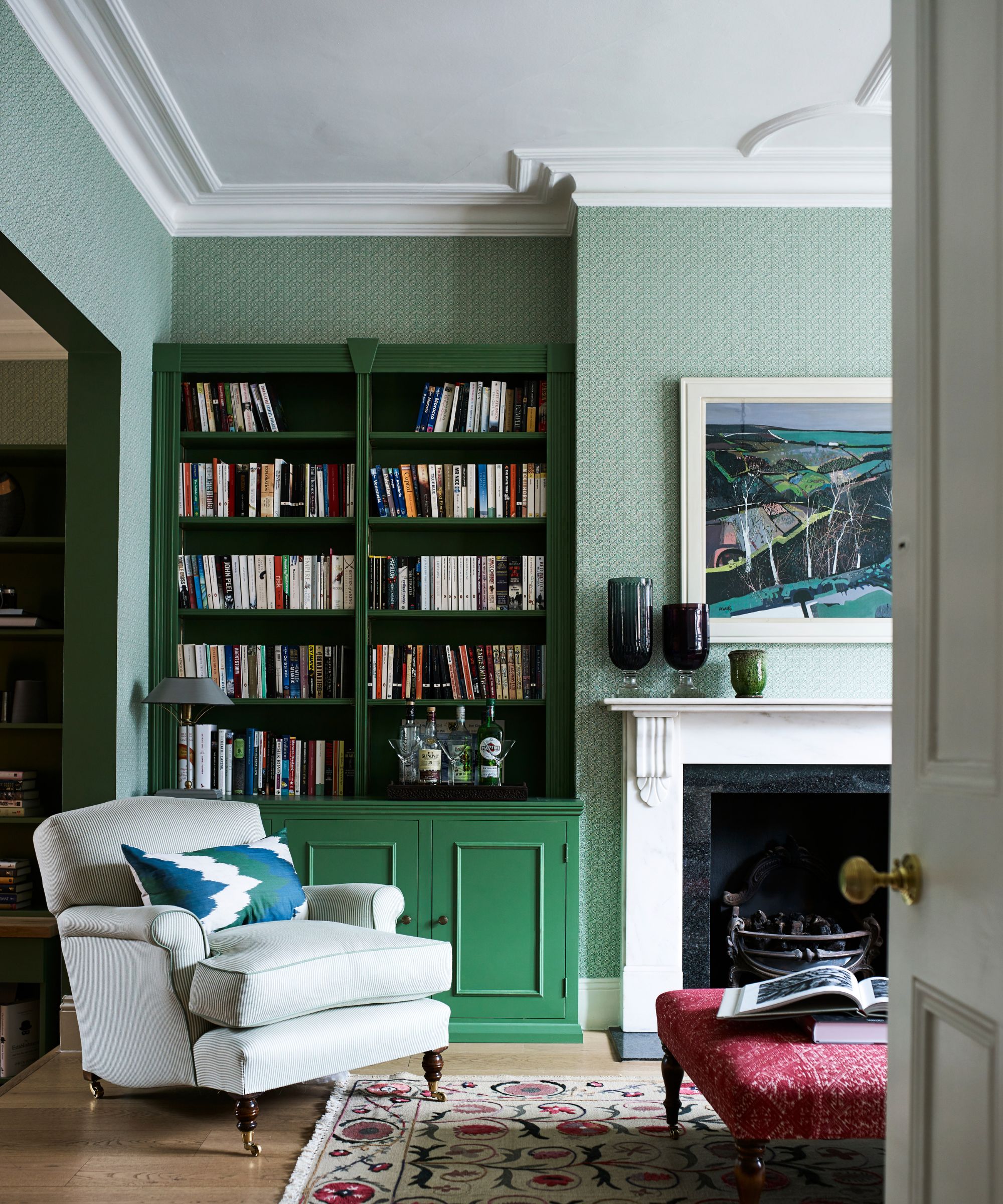 Green living room with bookshelf, cream armchair, fireplace with three vases and red ottoman