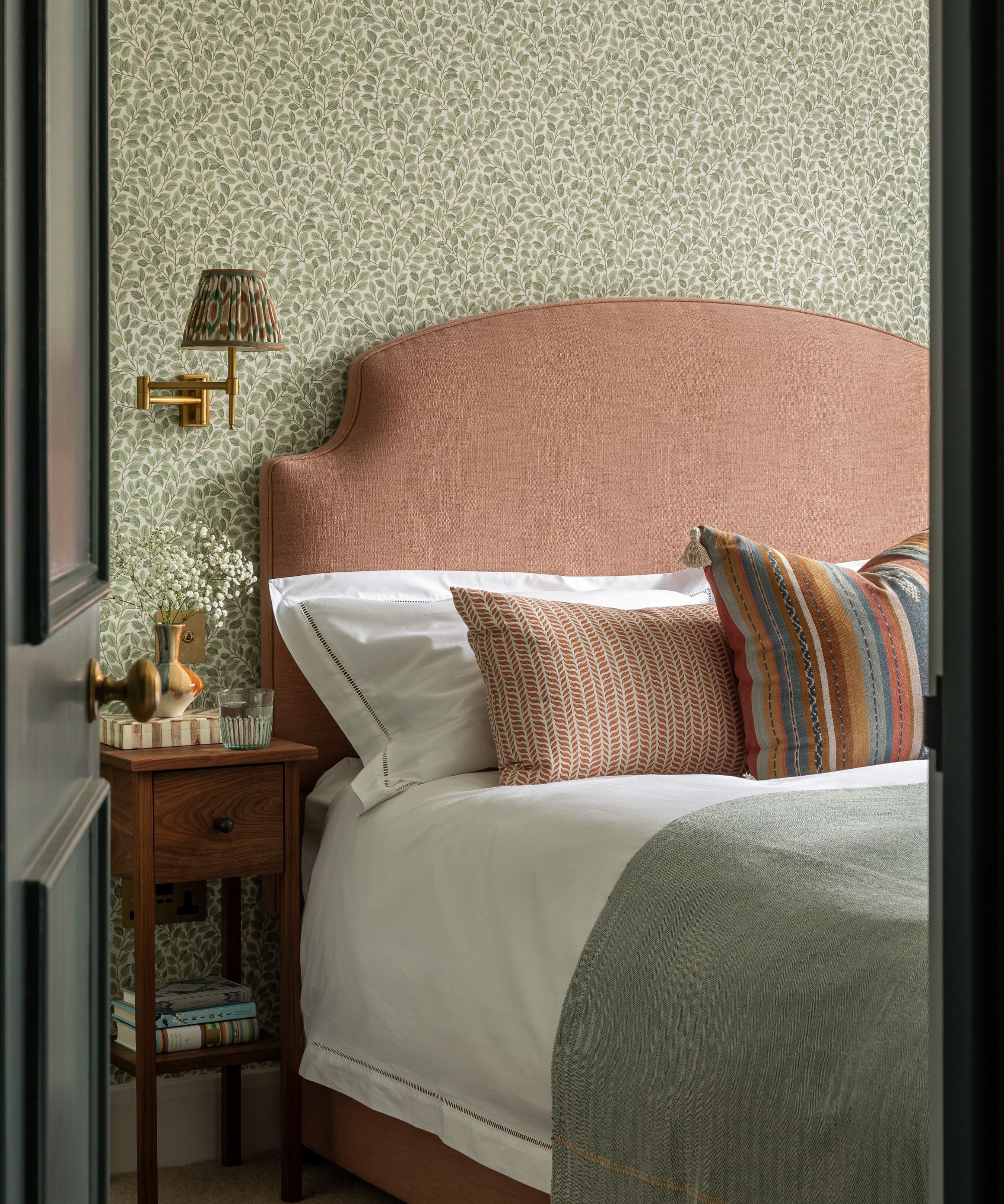 A bedroom with a large bed with a pastel orange headboard, and wallpaper with a green pattern