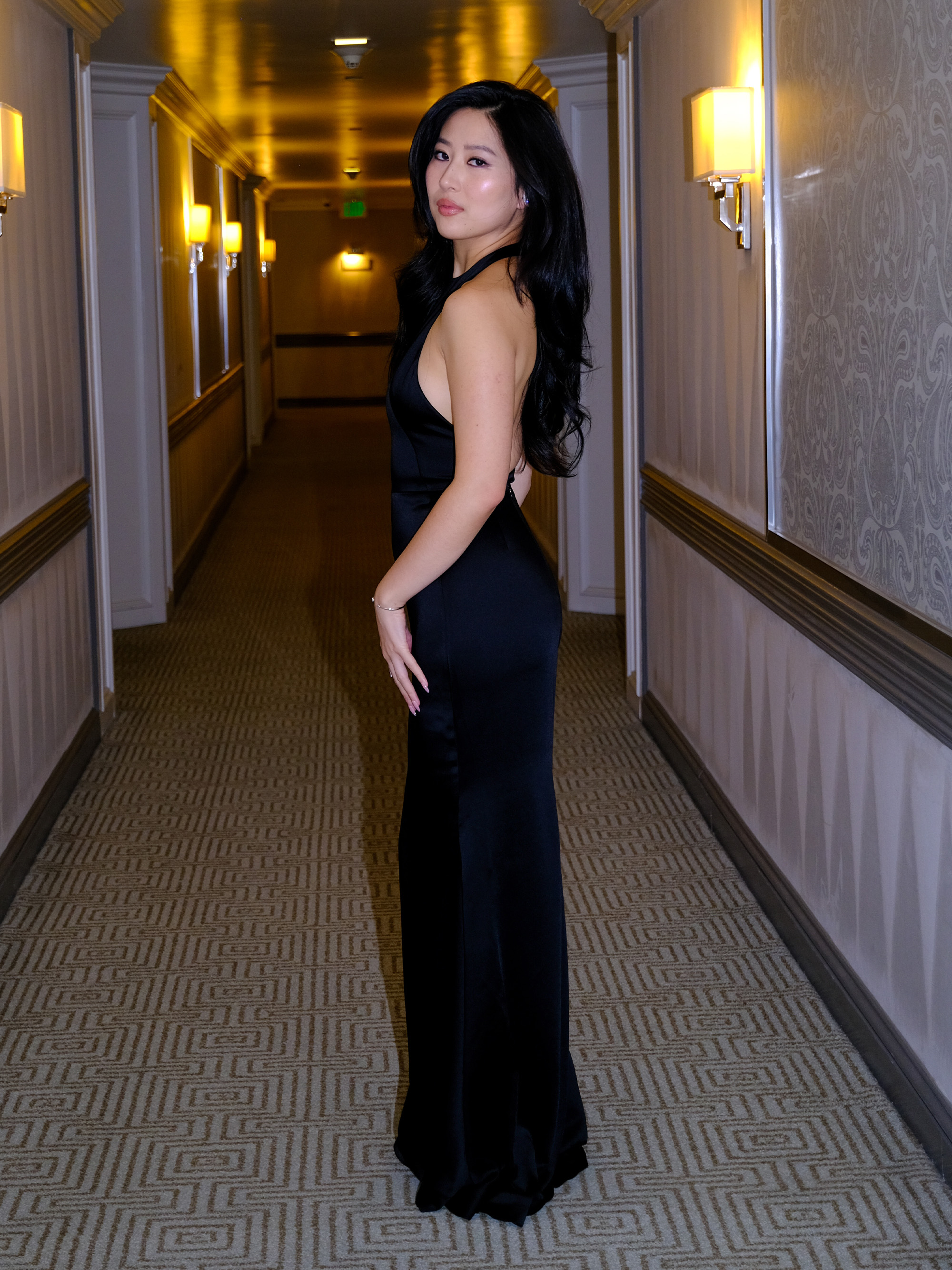 Mabel Li was photographed getting ready for the Los Angeles premiere of Testament. He is shot in the hallway of his hotel looking back at the camera. She is wearing a black Givenchy gown with a halter neck.