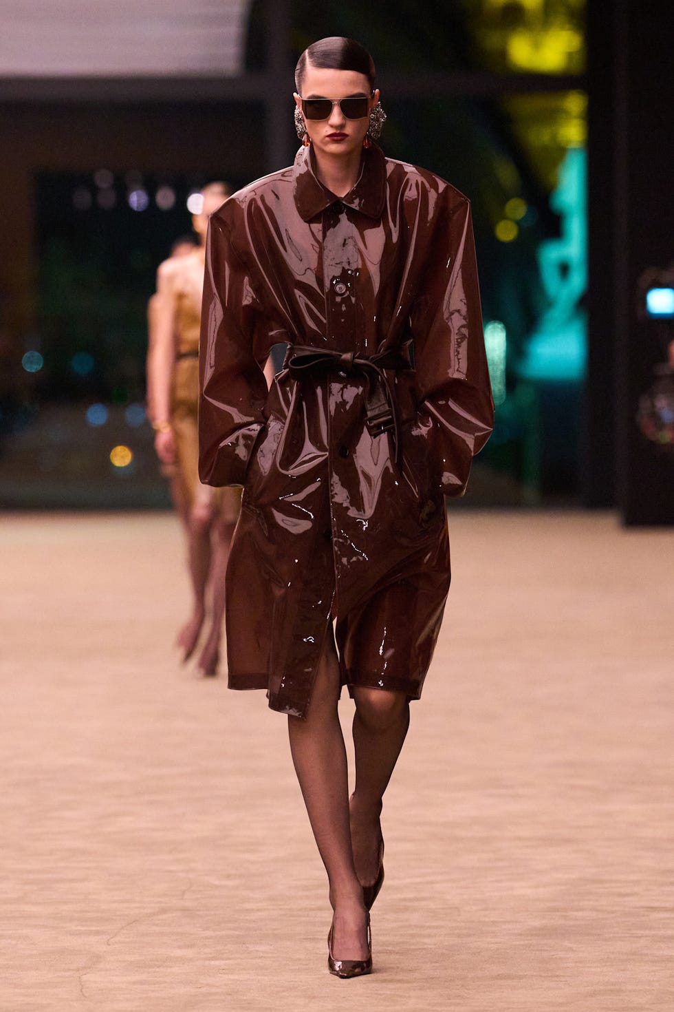 Model walking in a shiny brown coat on the runway.
