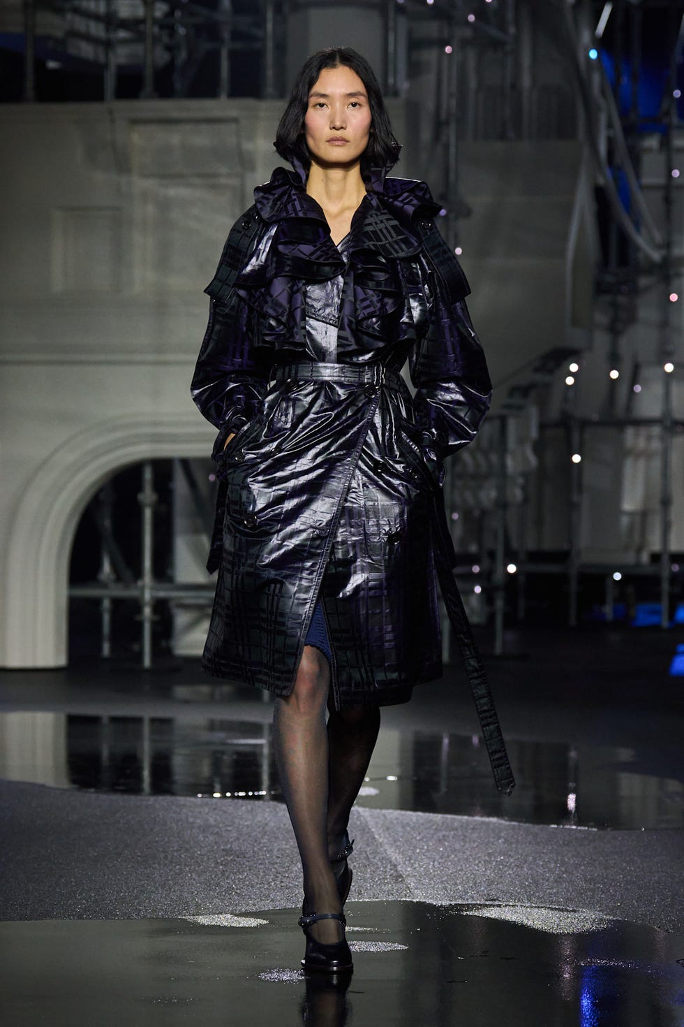 A model walking the runway is wearing a black, shiny trench coat.