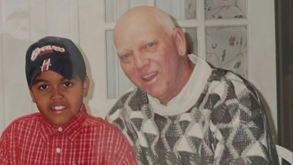 A young Druski in a red plaid shirt and Braves cap sits next to an older man in a patterned sweater, both smiling indoors.