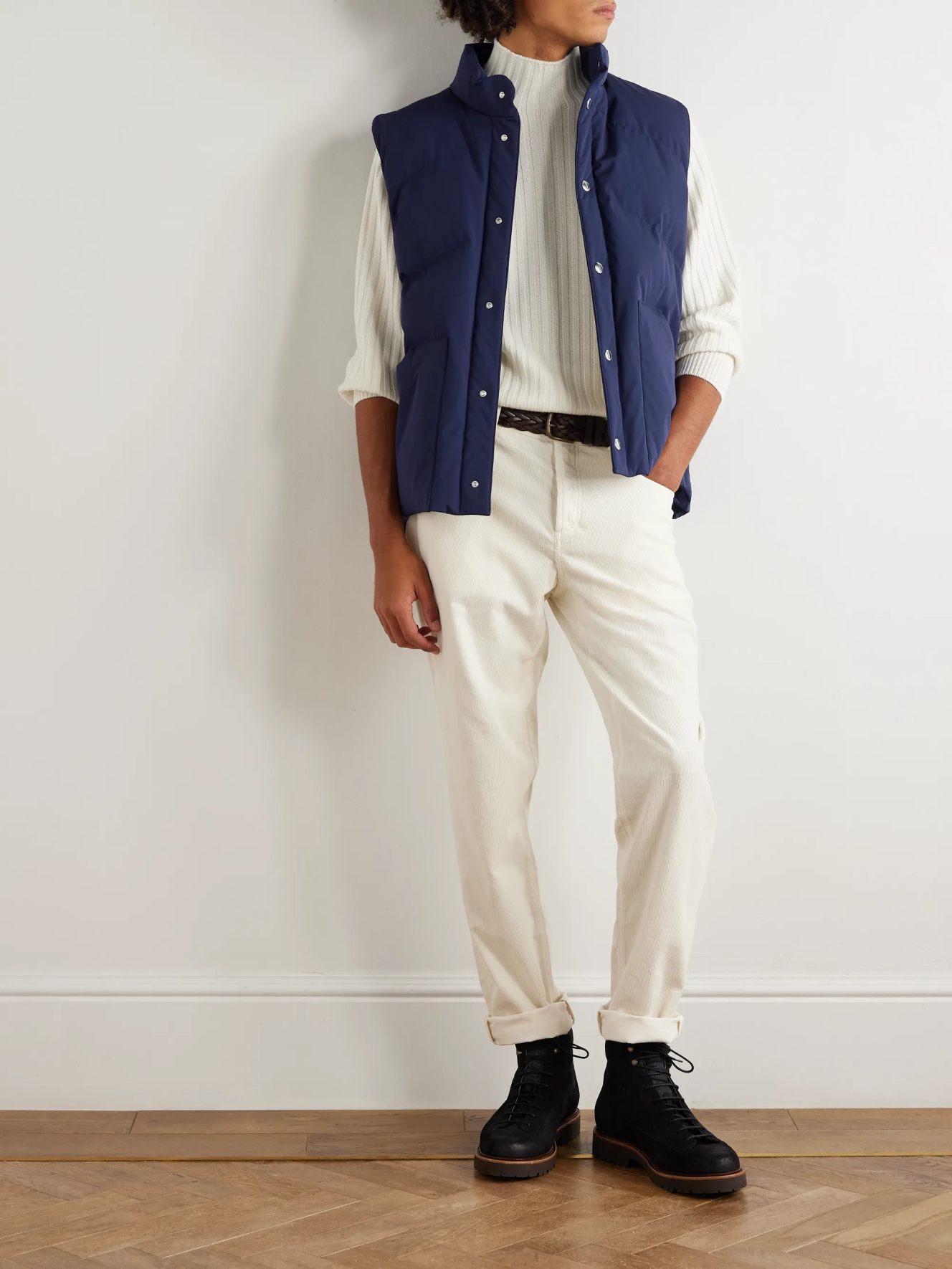 One models a light blue vest over a cream turtleneck, cream corduroy pants, and black boots with cuffed cuffs.