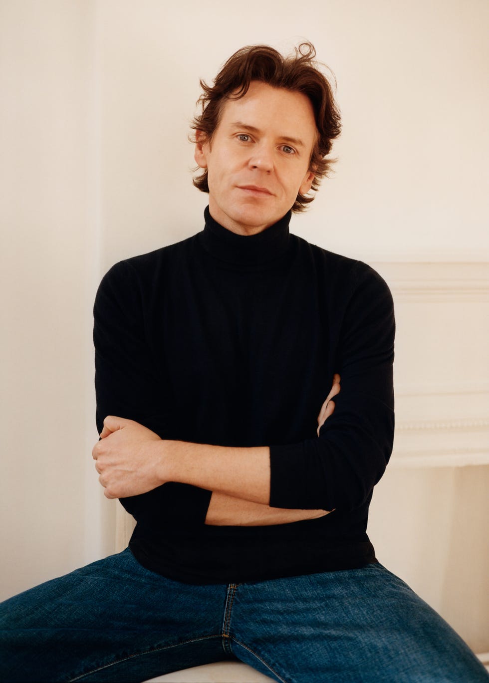 A person sitting with arms raised, wearing a black turtleneck and blue jeans.